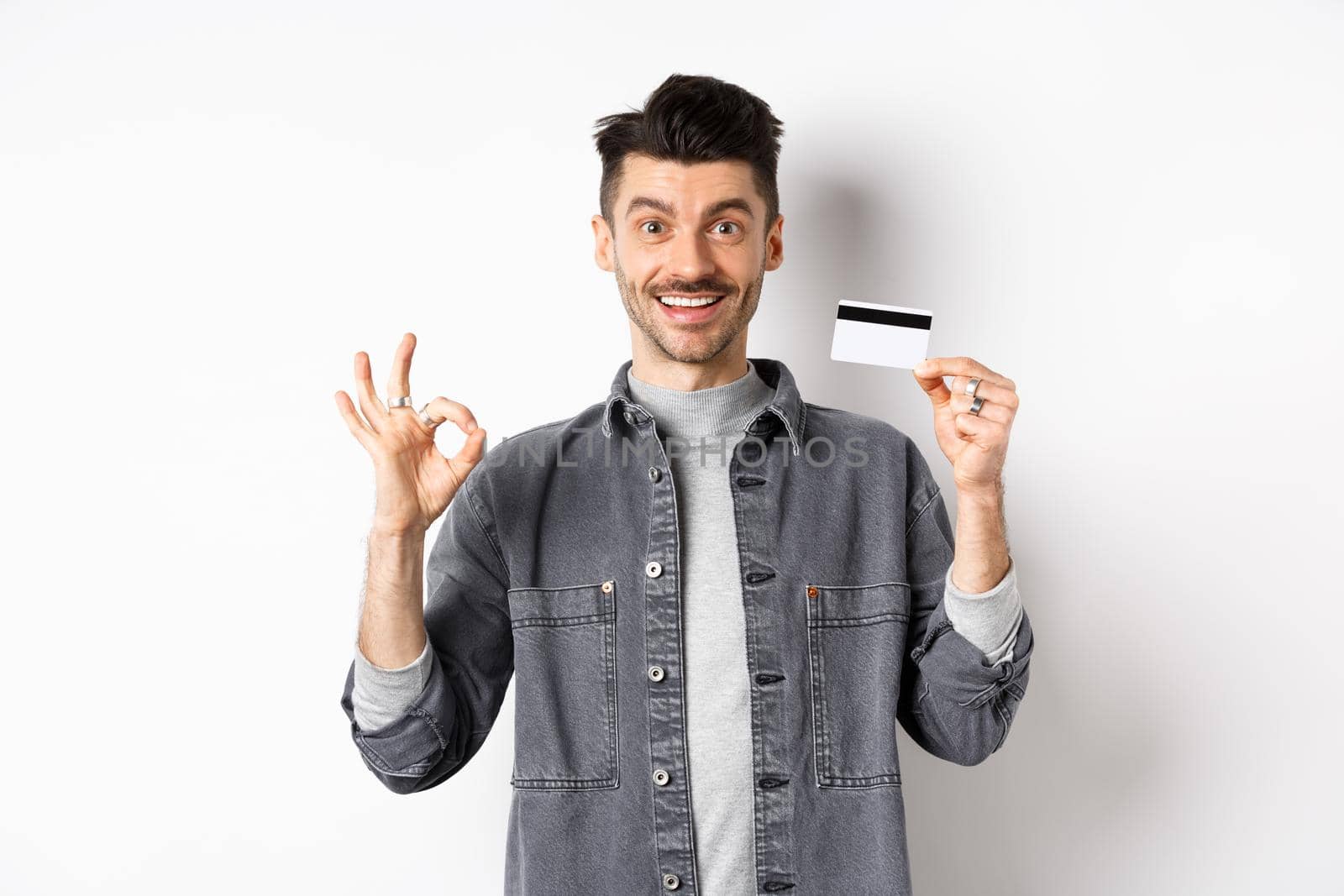 Very good. Smiling guy with plastic credit card showing okay sign, smiling satisfied, recommend bank, standing on white background.