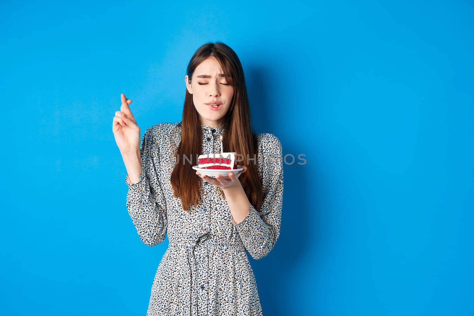 Hopeful birthday girl making wish on cake, cross fingers for good luck, standing in trendy dress against blue background by Benzoix