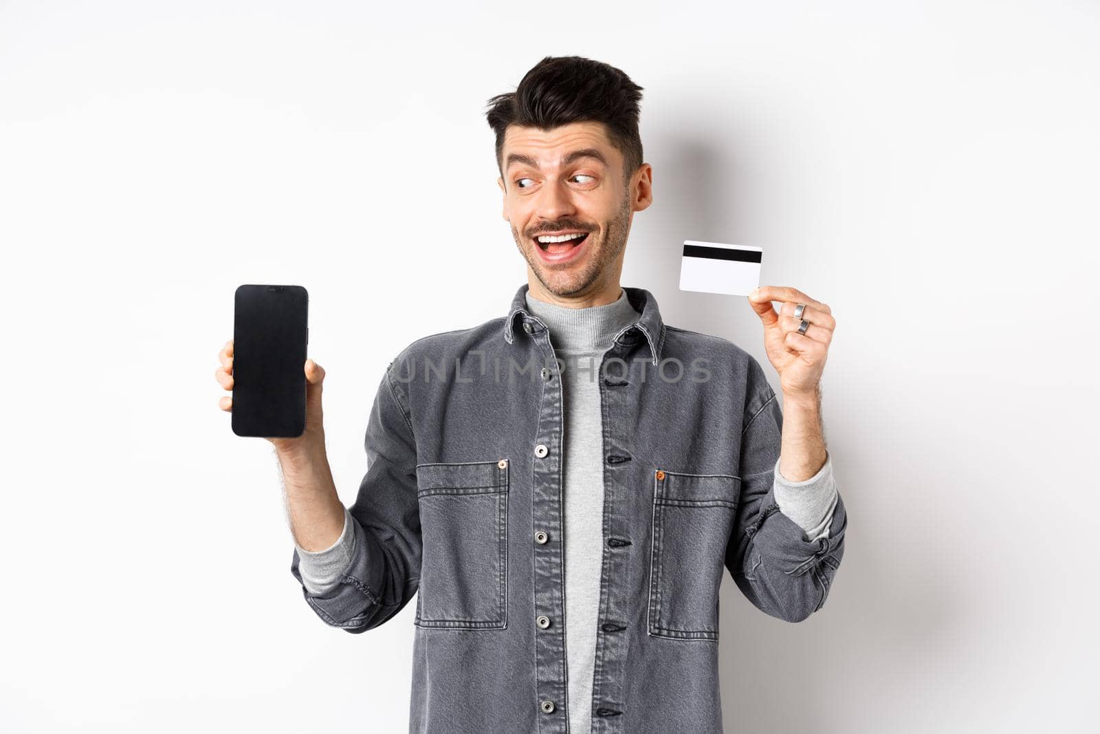 Handsome smiling man in denim jacket looking at empty smartphone screen and showing plastic credit card, good internet offer, standing against white background.