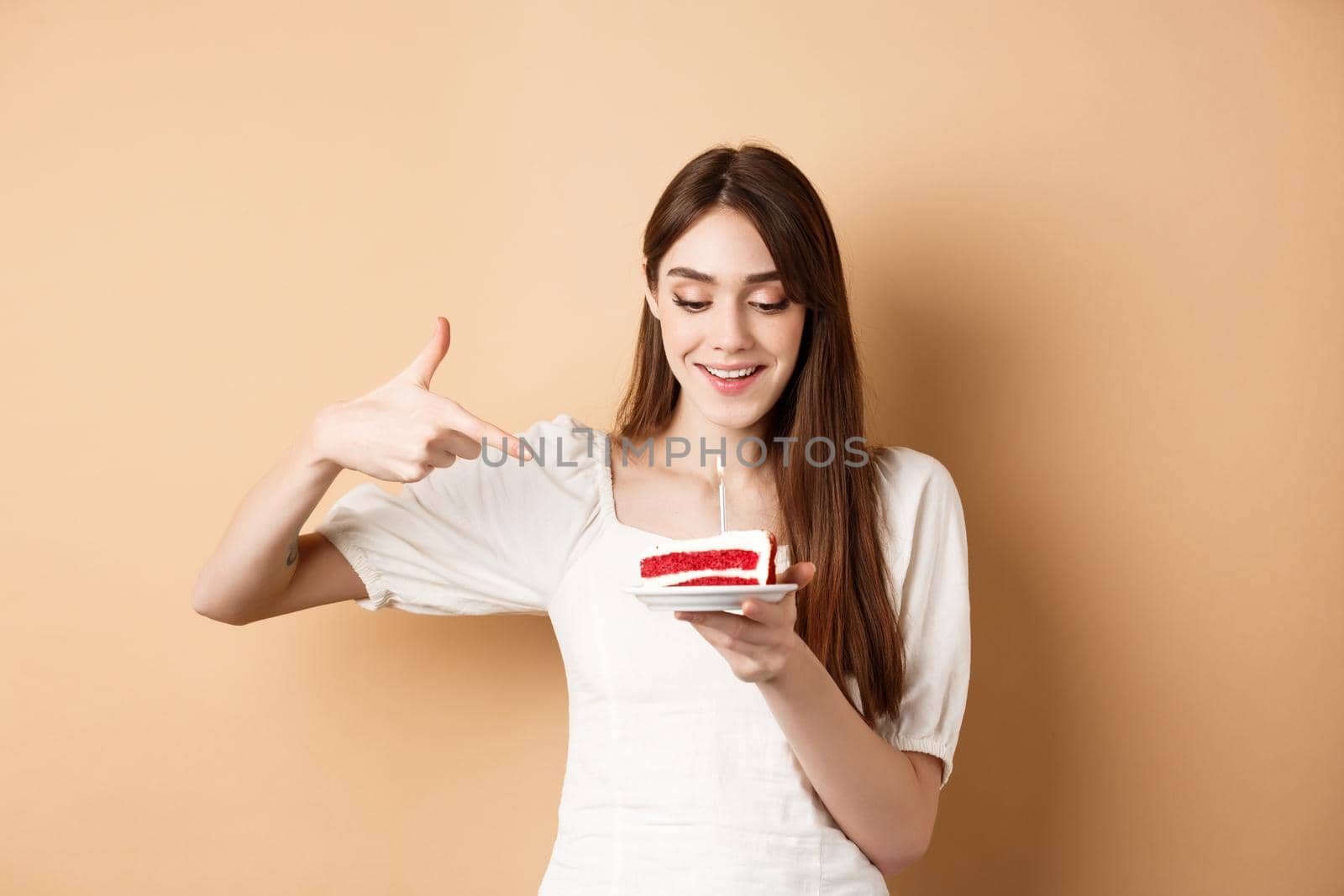 Cute girl pointing at birthday cake with candle and smiling excited, making bday wish, standing on beige background.