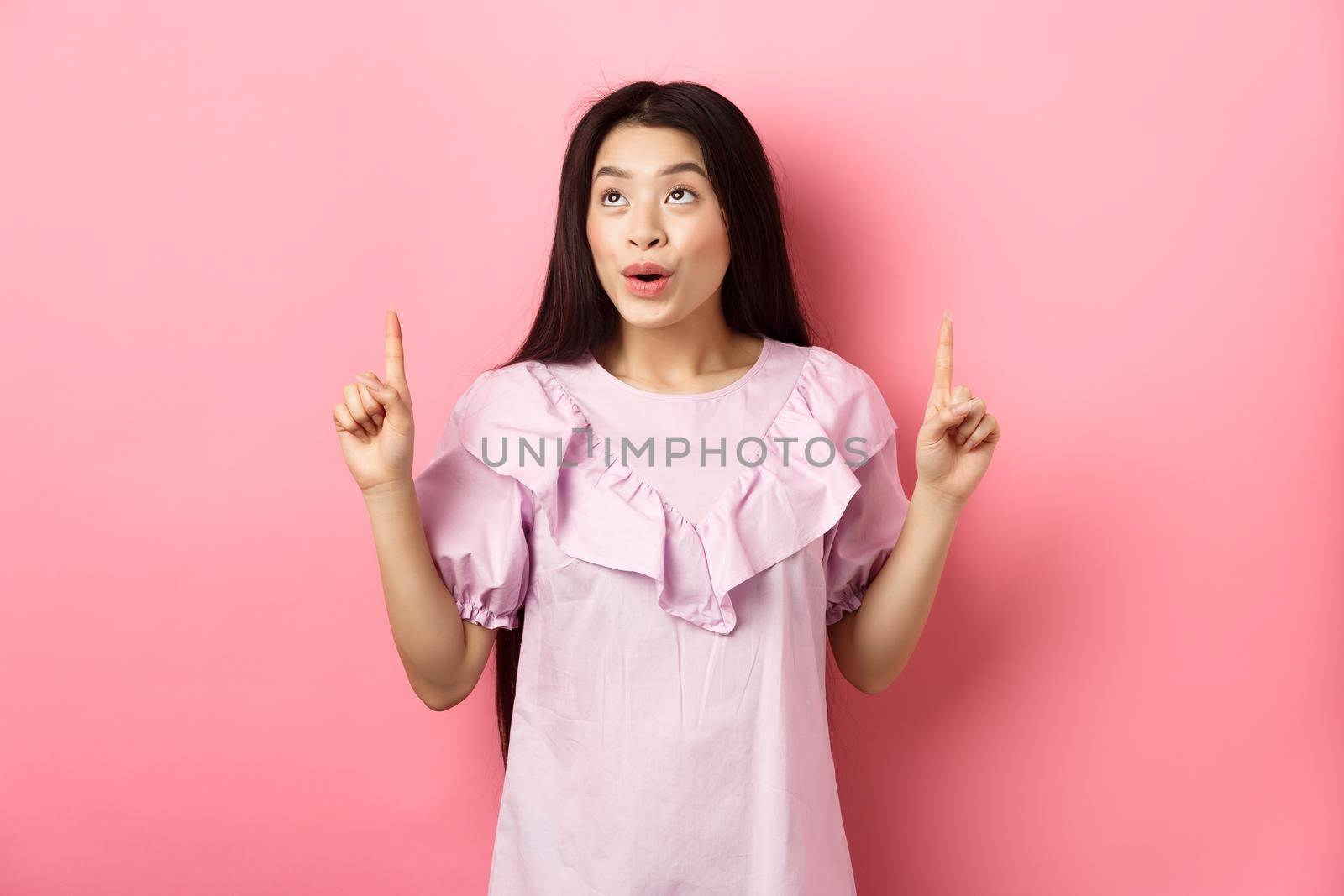 Excited teen asian woman pointing fingers up, looking dreamy at empty space, standing in dress against pink background by Benzoix