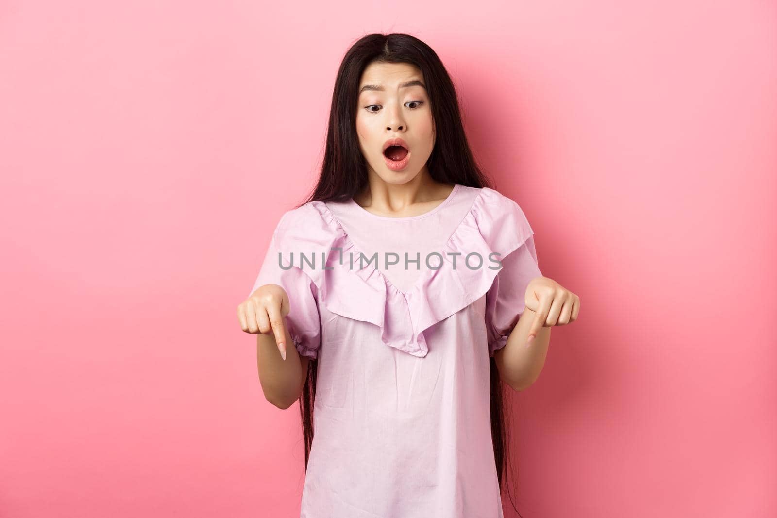 Surprised asian woman in awe, drop jaw and gasping, pointing fingers down at amazing promo offer, standing impressed against pink background by Benzoix