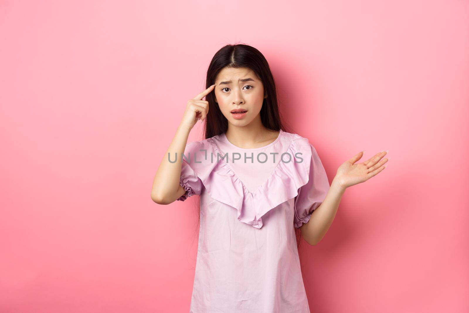 Are you crazy. Shocked asian woman pointing at head temple and complaining someone stupid action, scolding person, standing against pink background.