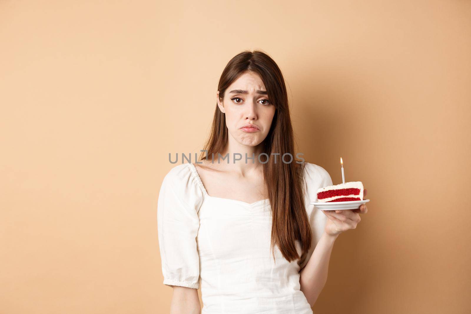 Sad birthday girl holding cake with candle and look upset, feeling lonely on her bday, standing alone on beige background by Benzoix