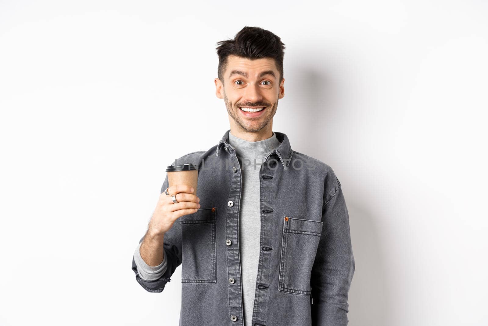 Enthusiastic handsome man holding coffee cup and smiling, drinking good drink from cafe takeout, standing against white background.