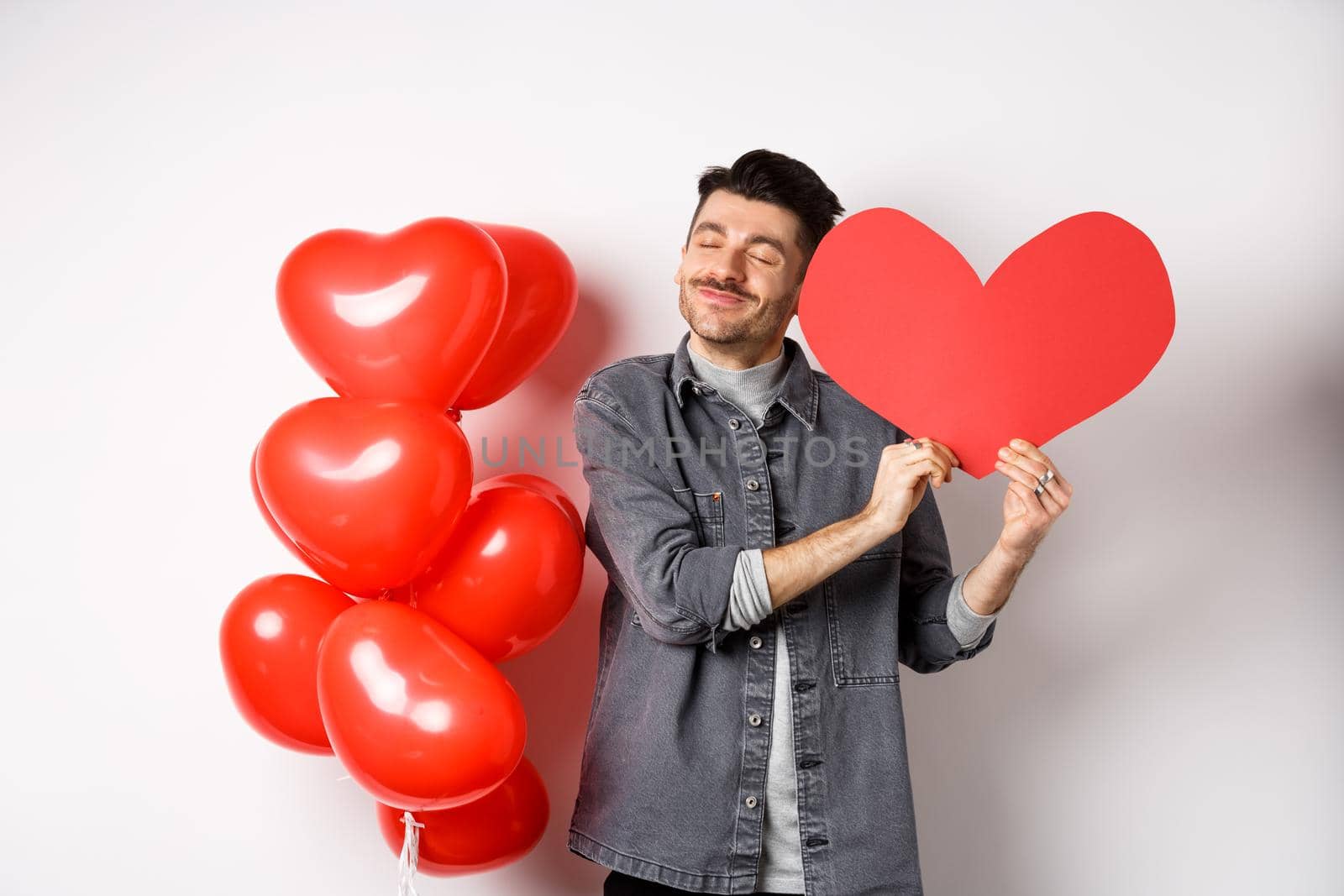 Romantic guy celebrating Valentines day, hugging big red heart card from lover and smiling happy, being in love, standing on white background near red balloons.