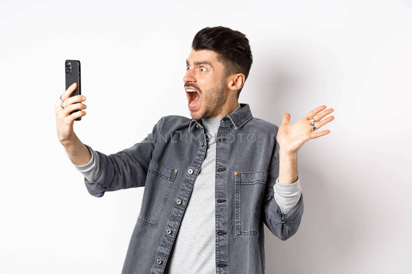 Shocked and confused man staring frustrated at smartphone screen, shouting from disappointment, standing against white background.