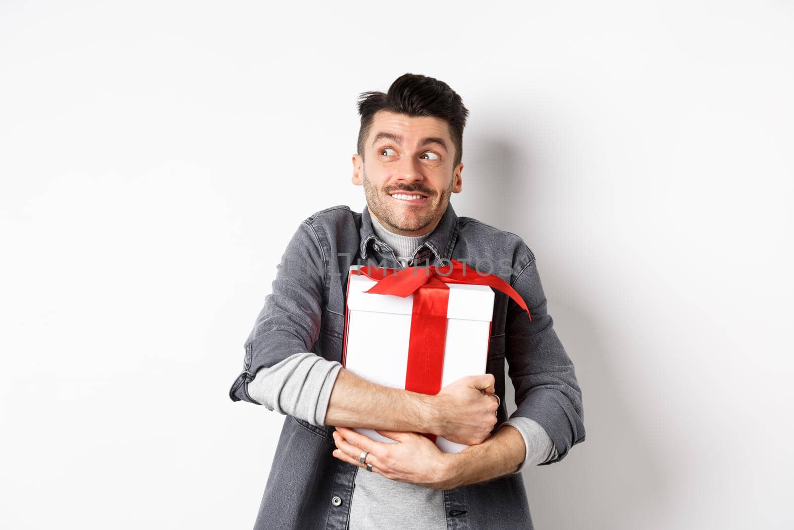 Happy boyfriend hugging gift from lover, looking aside at empty space and smiling excited, making romantic present on valentines day, standing on white background.