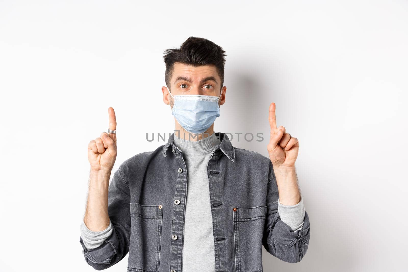 Pandemic lifestyle, healthcare and medicine concept. Stylish modern man in medical mask showing advertisement, pointing fingers up at logo, standing on white background by Benzoix