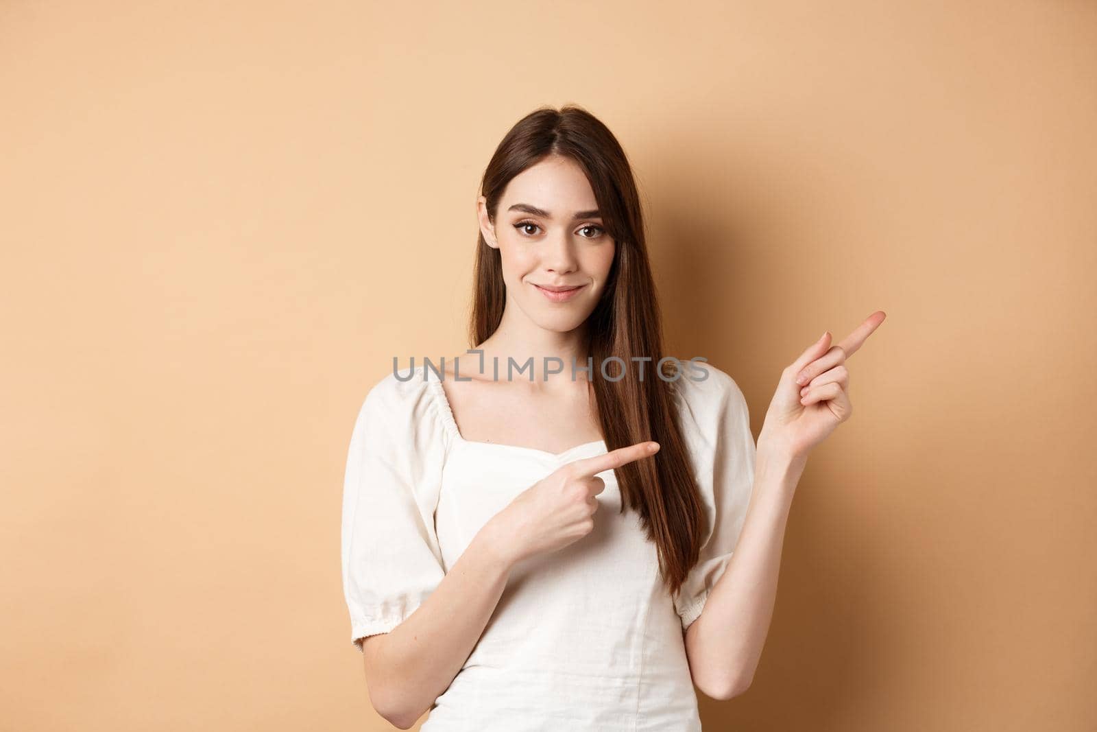 Beautiful young woman in dress pointing fingers right, showing logo and looking tender at camera, beige background by Benzoix