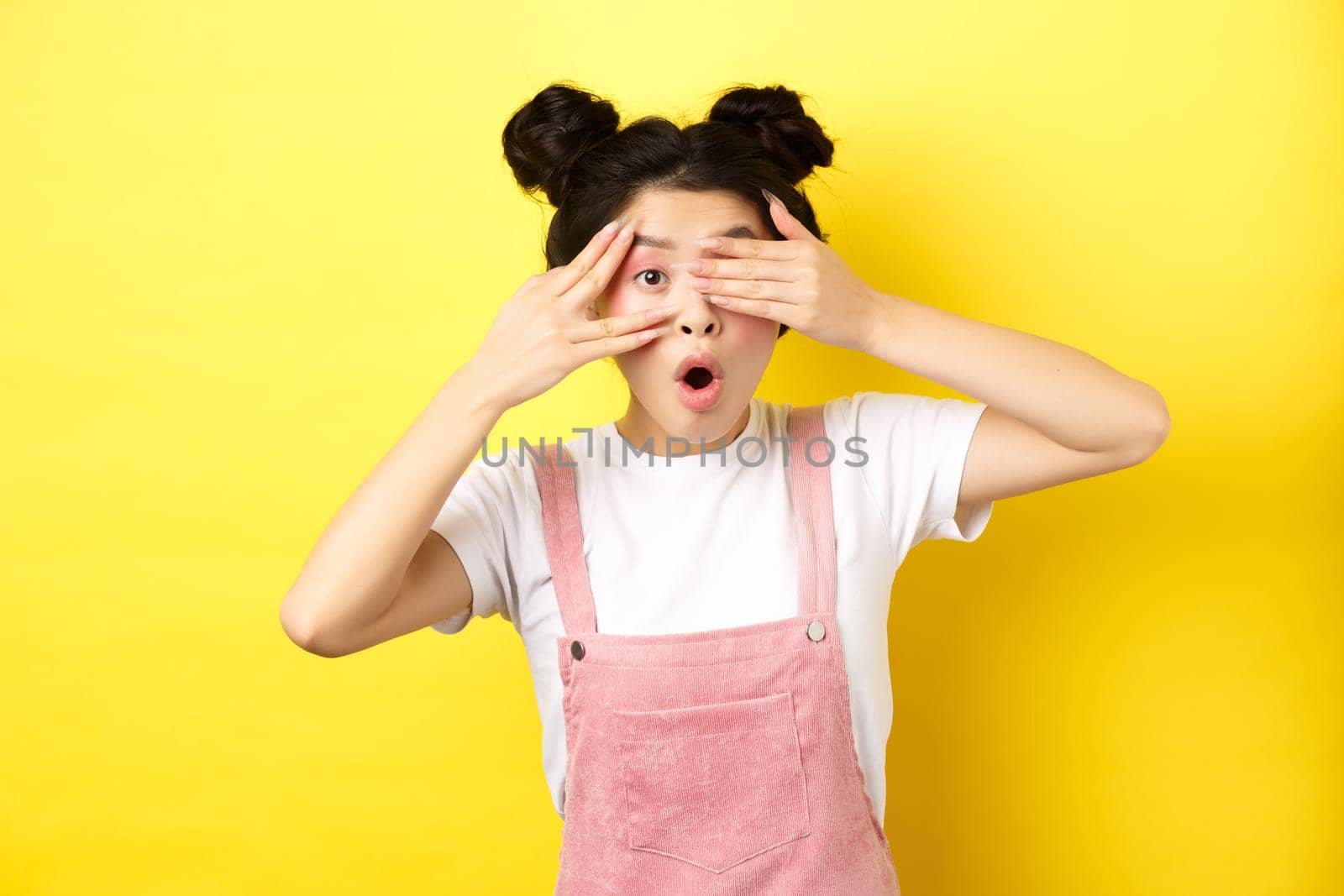 Excited asian teen girl covering eyes with hands and peeking through fingers, gasping amazed at camera, standing in summer clothes on yellow background by Benzoix