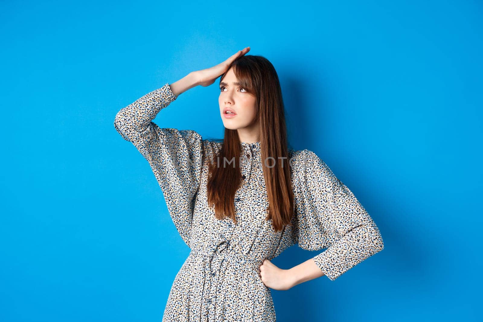 Troubled young woman in dress slap forehead and roll eyes up, standing tired and bothered with work, blue background.