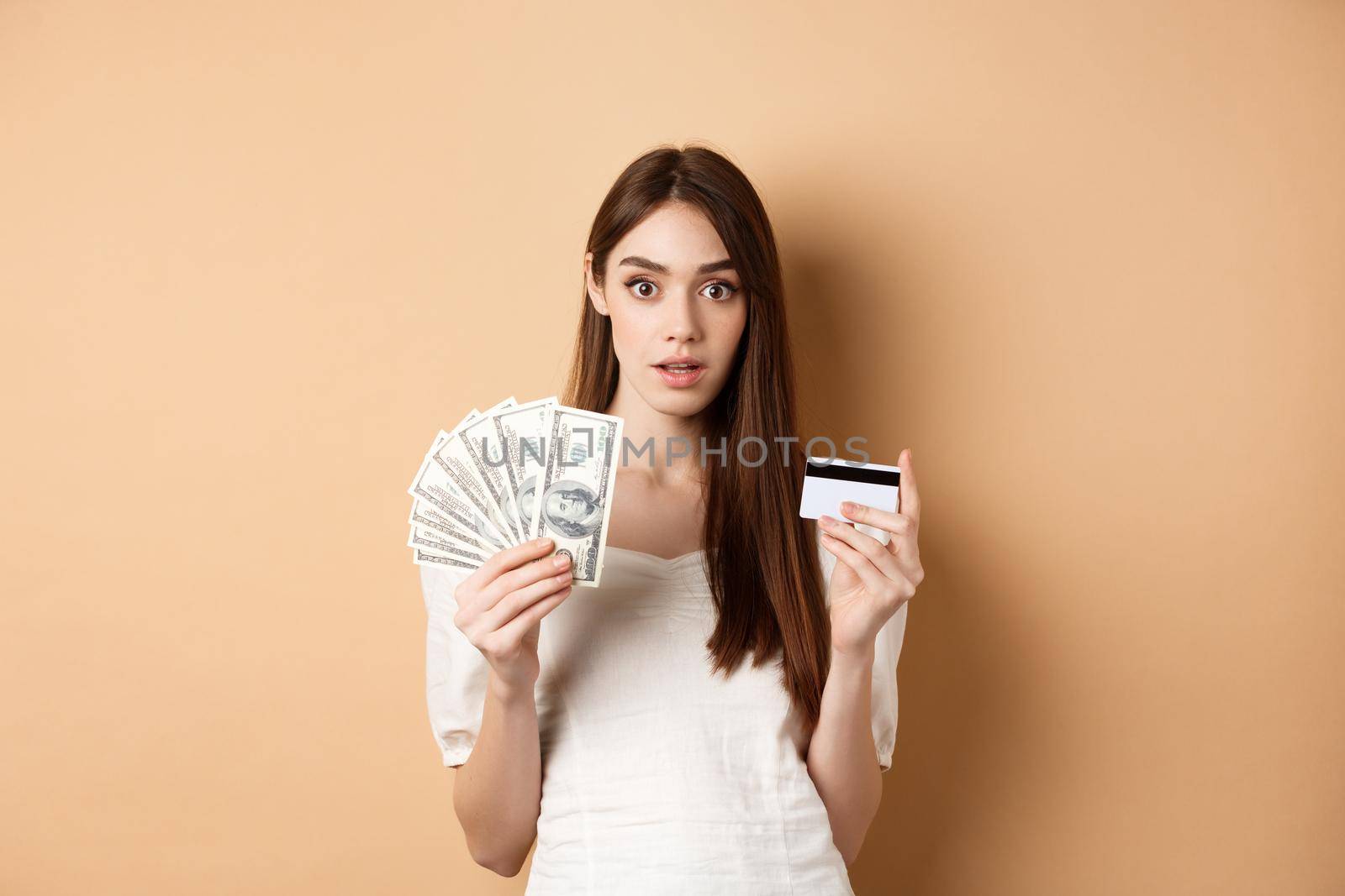 Excited woman holding dollar bills and plastic credit card, looking amazed at camera, standing on beige background.