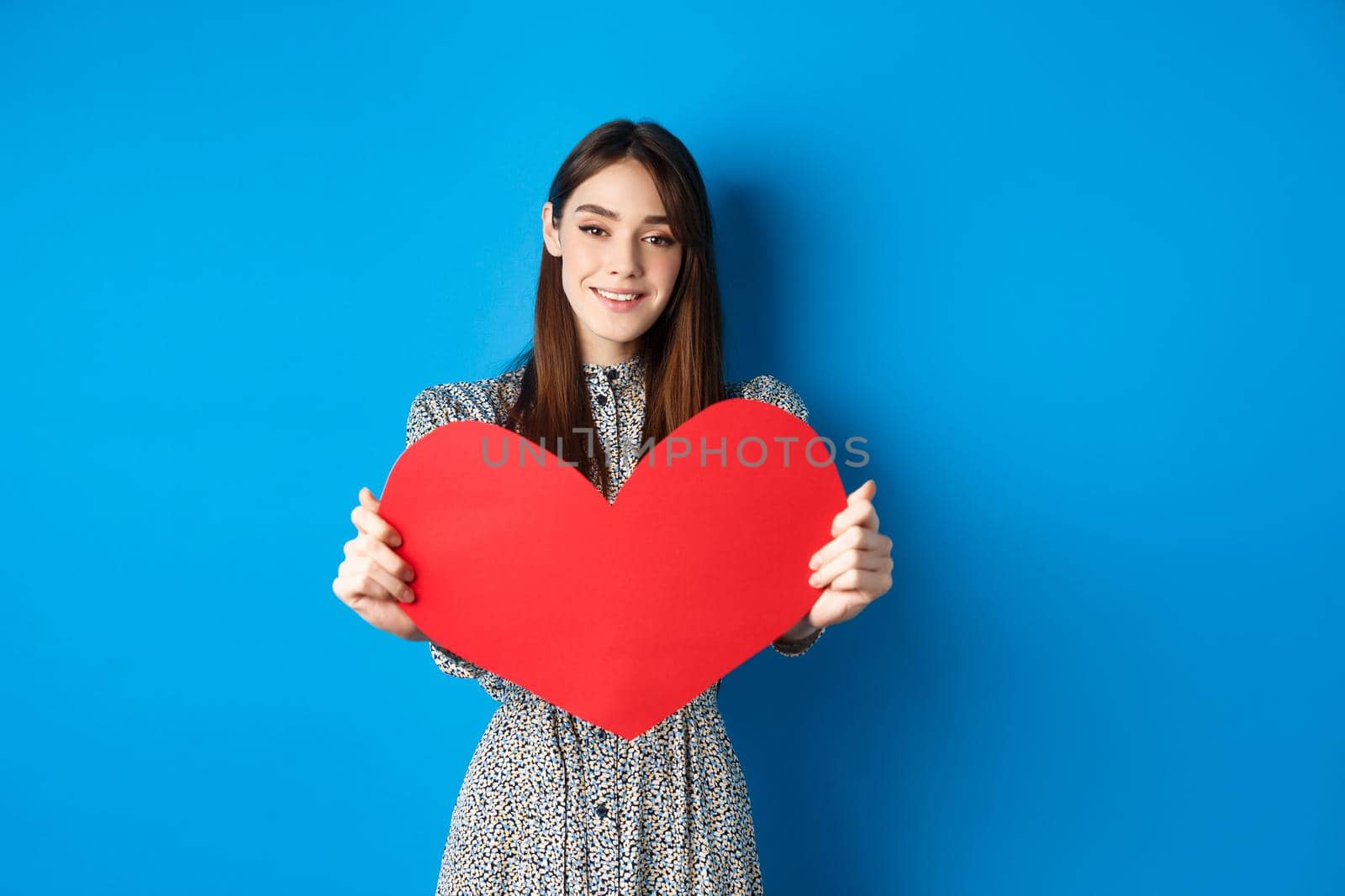 Valentines day and relationship concept. Beautiful romantic girl say I love you, extending hand with big red heart, standing on blue background by Benzoix