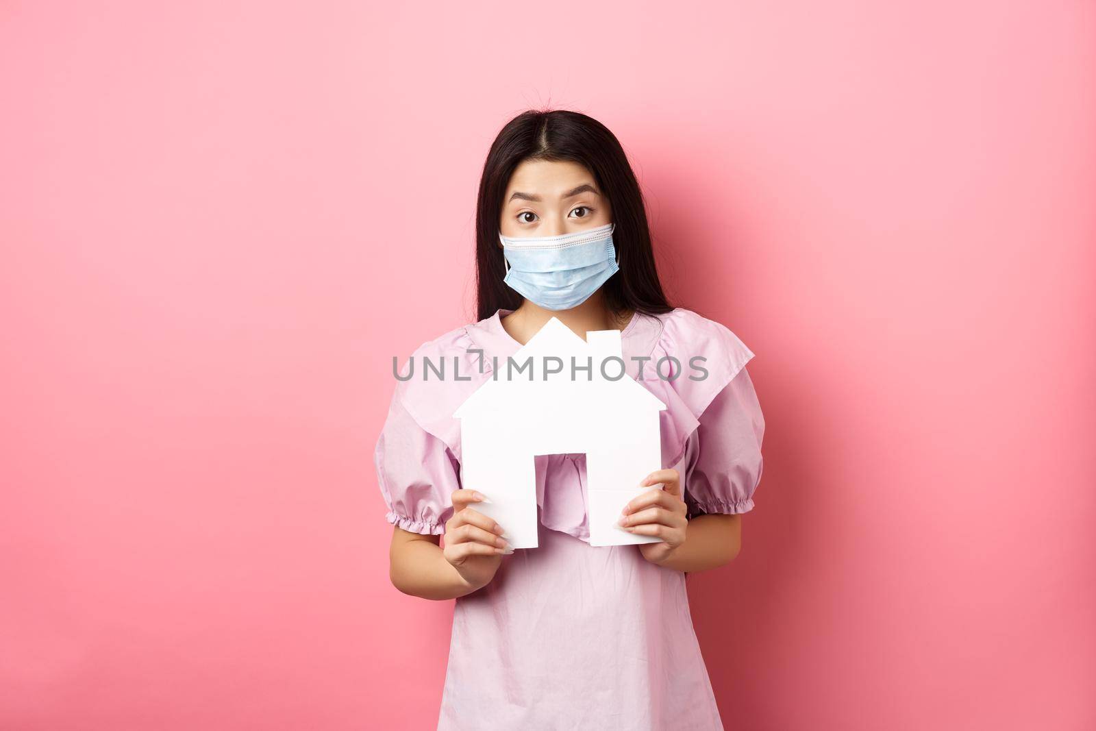 Real estate and pandemic concept. Excited asian girl in medical mask showing paper house cutout, standing against pink background.