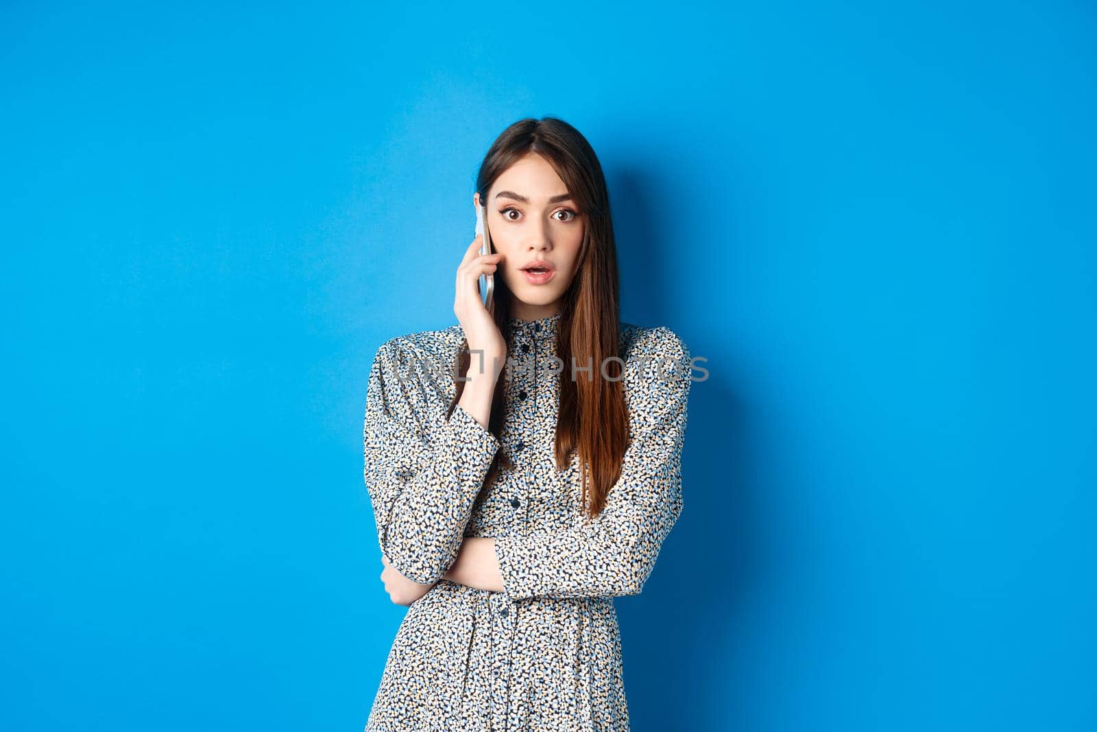 Shocked girl talking on mobile phone, gasping amazed at camera, speaking with someone and hear shocking news, standing in dress on blue background by Benzoix