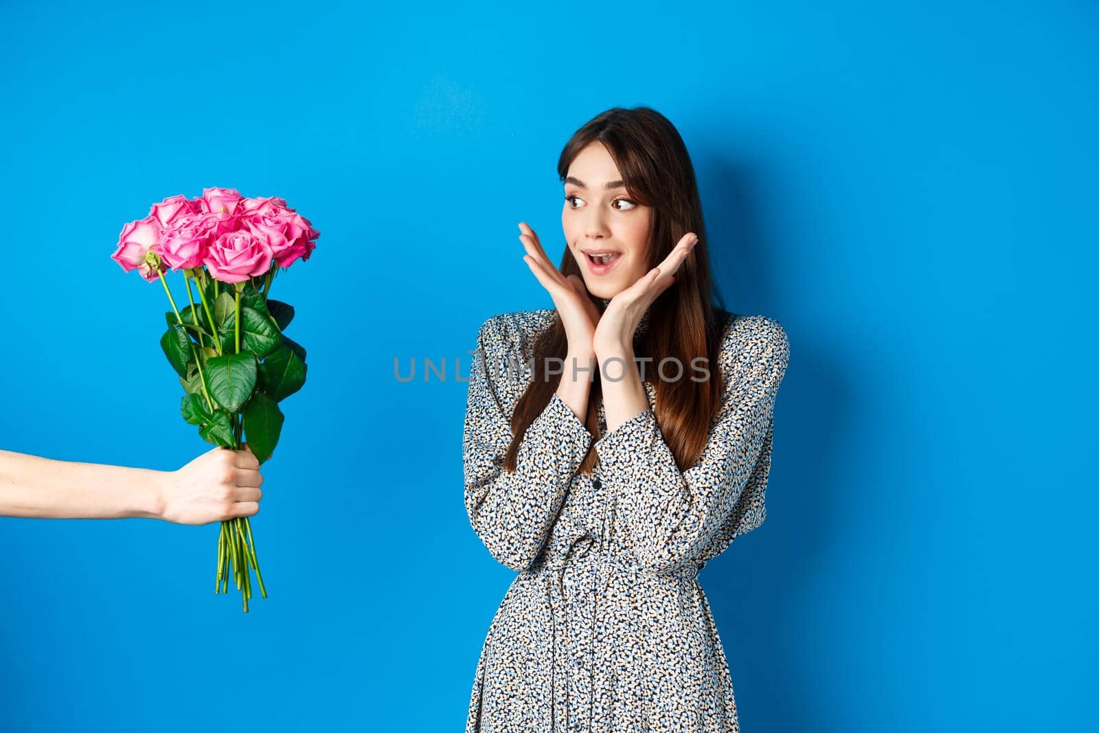 Valentines day concept. Romantic girl looking surprised at hand with flowers, woman receiving bouquet of pink roses, standing on blue background.