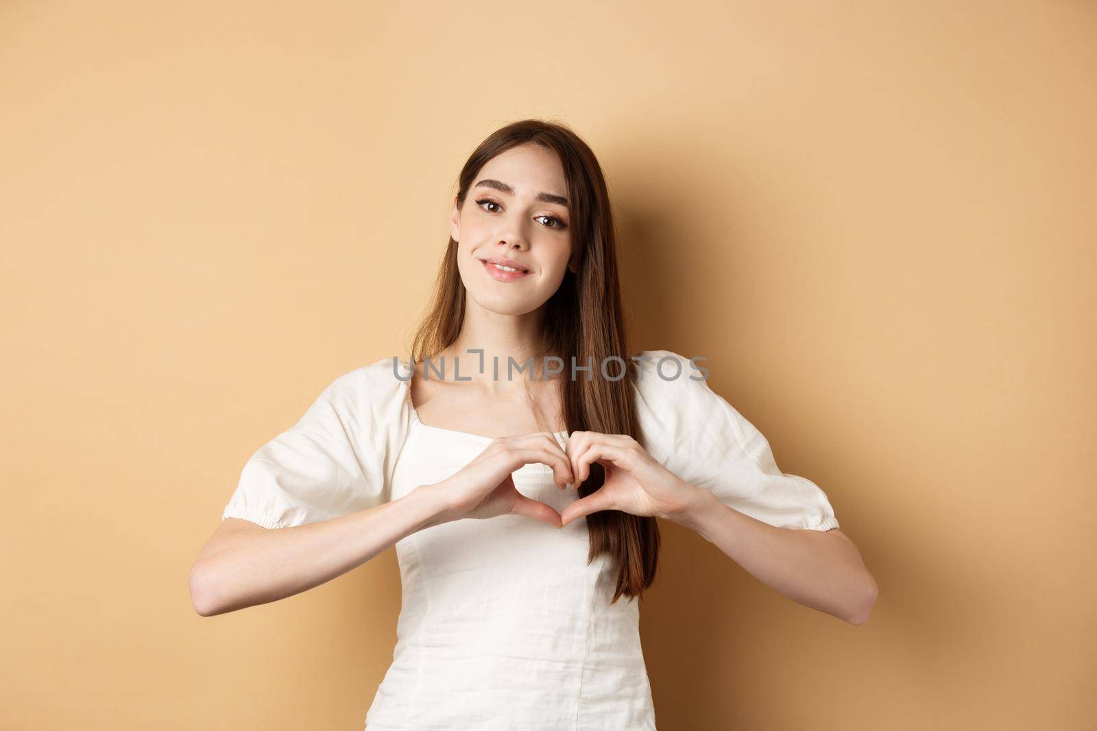 Beautiful caucasian woman say I love you, showing heart gesture and smile at camera, beige background. Concept of romance and Valentines day.