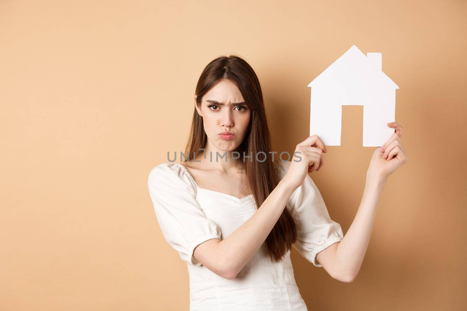 Real estate. Angry girl showing house cutout and frowning upset, complaining on apartment, standing against beige background.