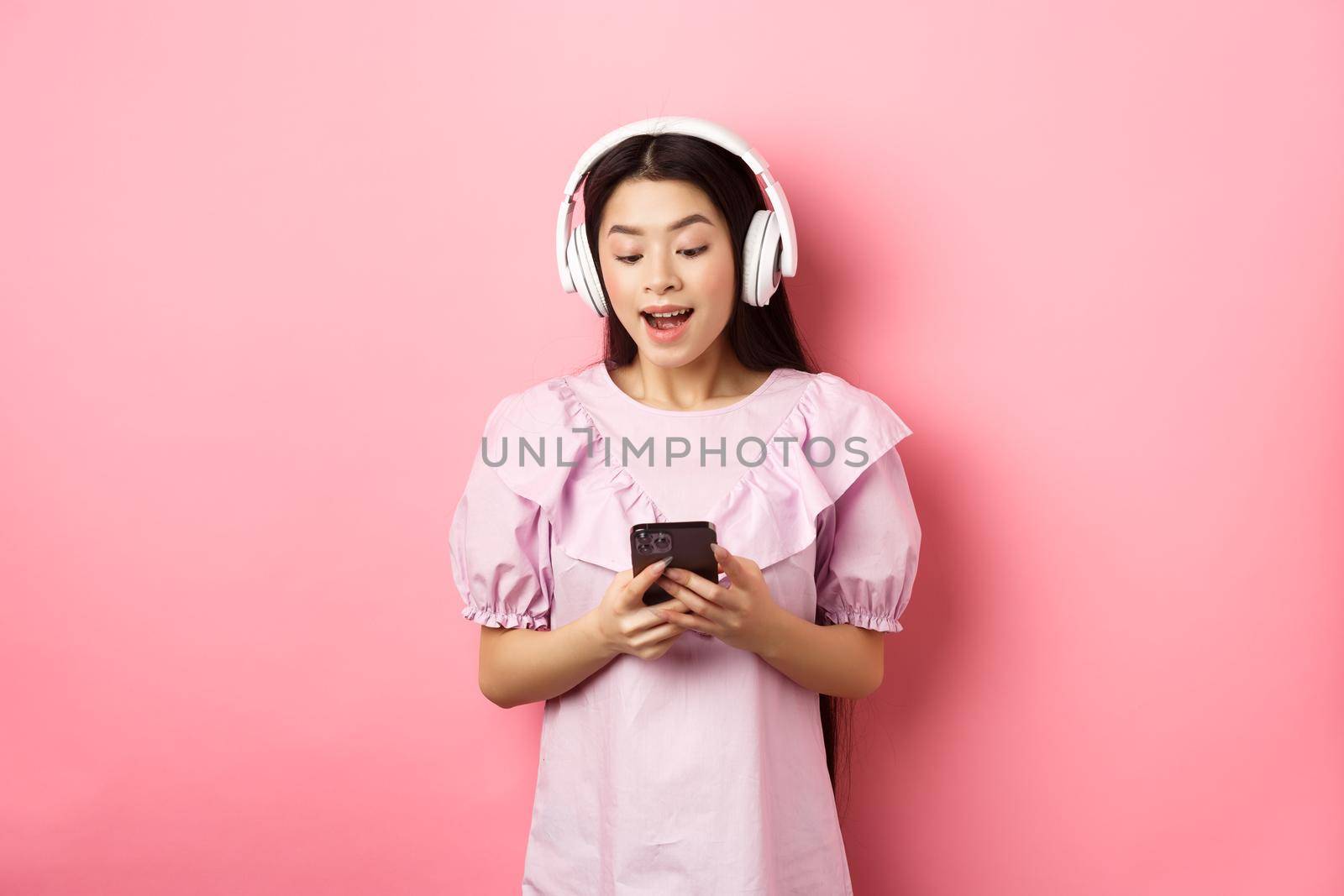 Happy beautiful woman in wireless headphones watching video on smartphone, looking at phone amused, standing against pink background by Benzoix