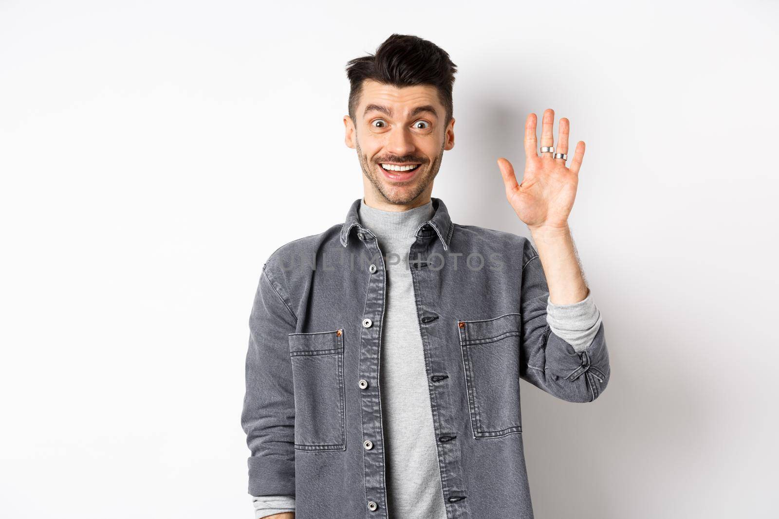 Friendly stylish man saying hello and waiving hand, smiling cheerful, greeting you with hi gesture, standing on white background.