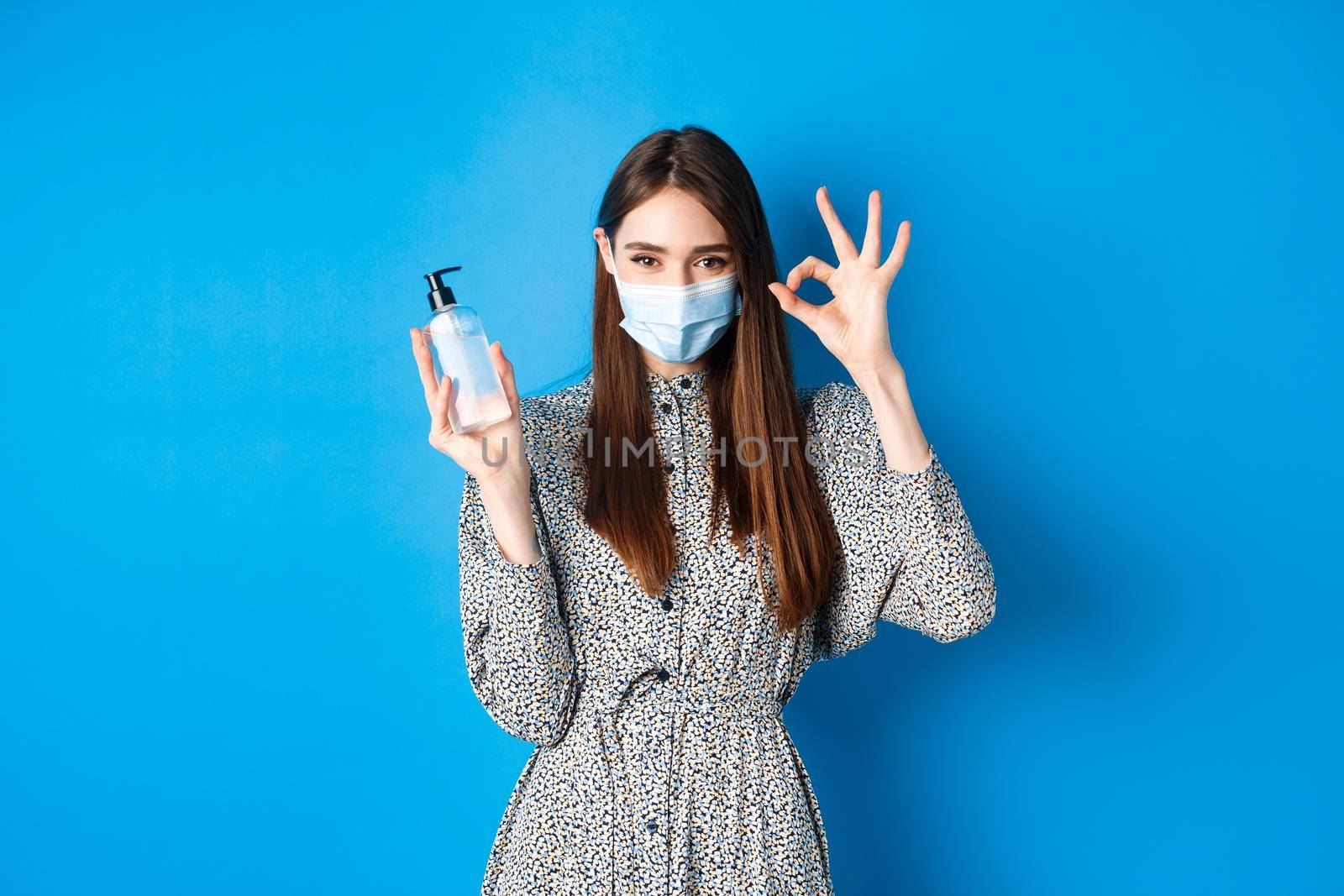 Covid-19, social distancing and healthcare concept. Beautiful girl in medical mask showing bottle antiseptic or hand sanitizer, make okay sign, recommend product, standing on blue background.