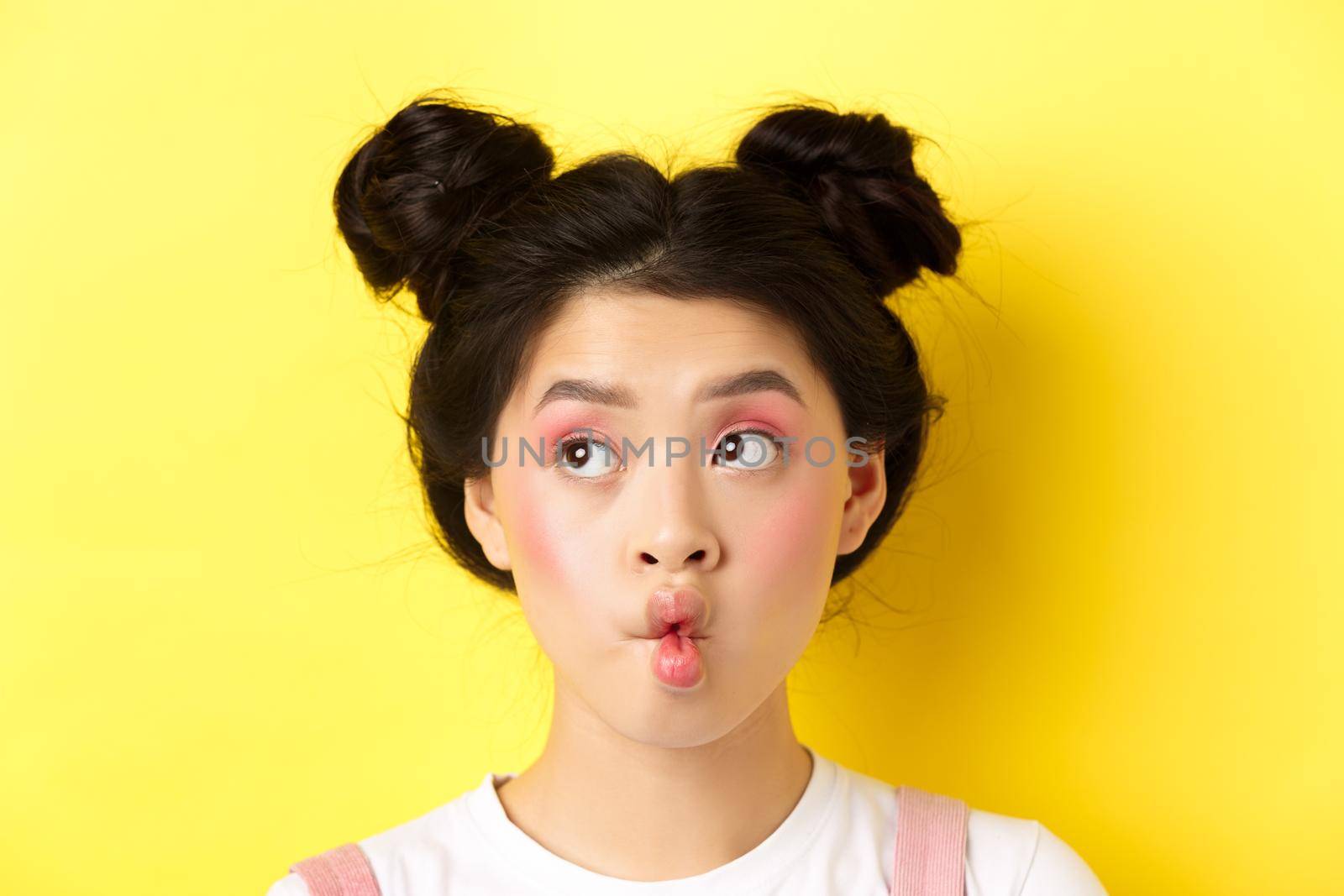 Close-up of teen asian girl pucker lips and looking funny at camera, standing with glamour makeup and stylish hairstyle, yellow background by Benzoix