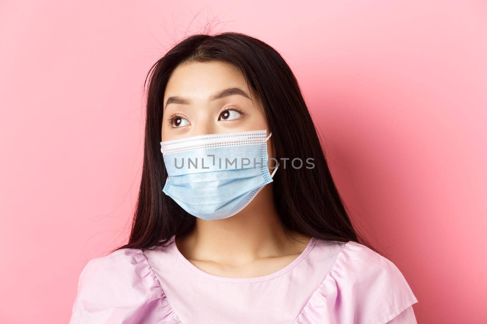 Covid-19, pandemic and quarantine concept. Close-up of dreamy asian girl in medical mask looking left at logo with thoughtful face, standing on pink background.