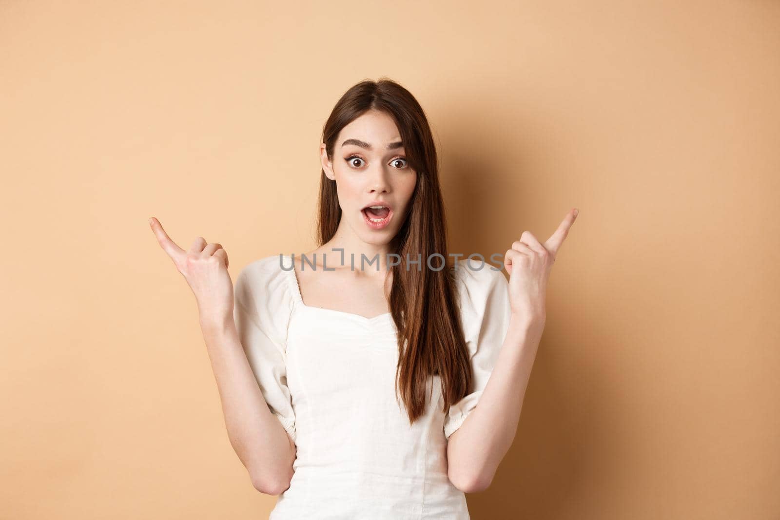 Surprised young girl pointing sideways at left and right, gasping and saying wow amazed, showing two cool choices, beige background.