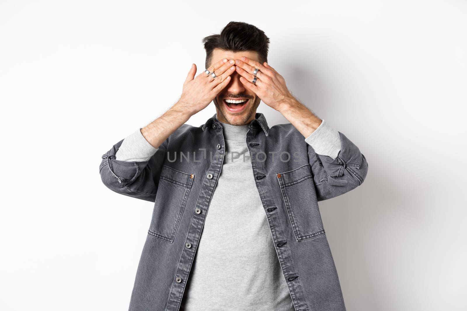 Handsome happy man cover his eyes and waiting for surprise, smiling excited, standing in casual clothes on white background.