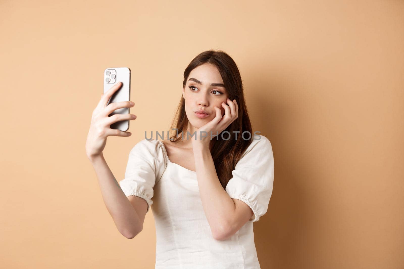 Stylish girl taking selfie, posing for mobile camera with photo filters, making cute face, standing on beige background by Benzoix