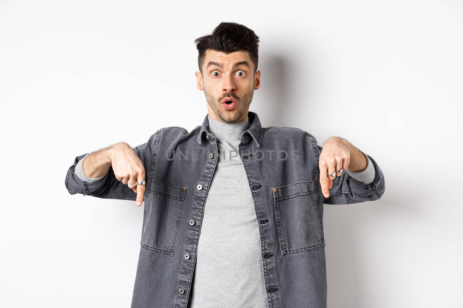 Excited guy say wow, pointing fingers down amazed, showing cool promo offer, standing on white background in denim jacket.