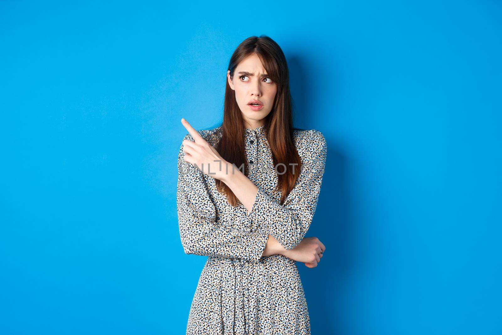 Confused girl in dress pointing and looking at upper left corner with puzzled frowning face, standing hesitant on blue background.