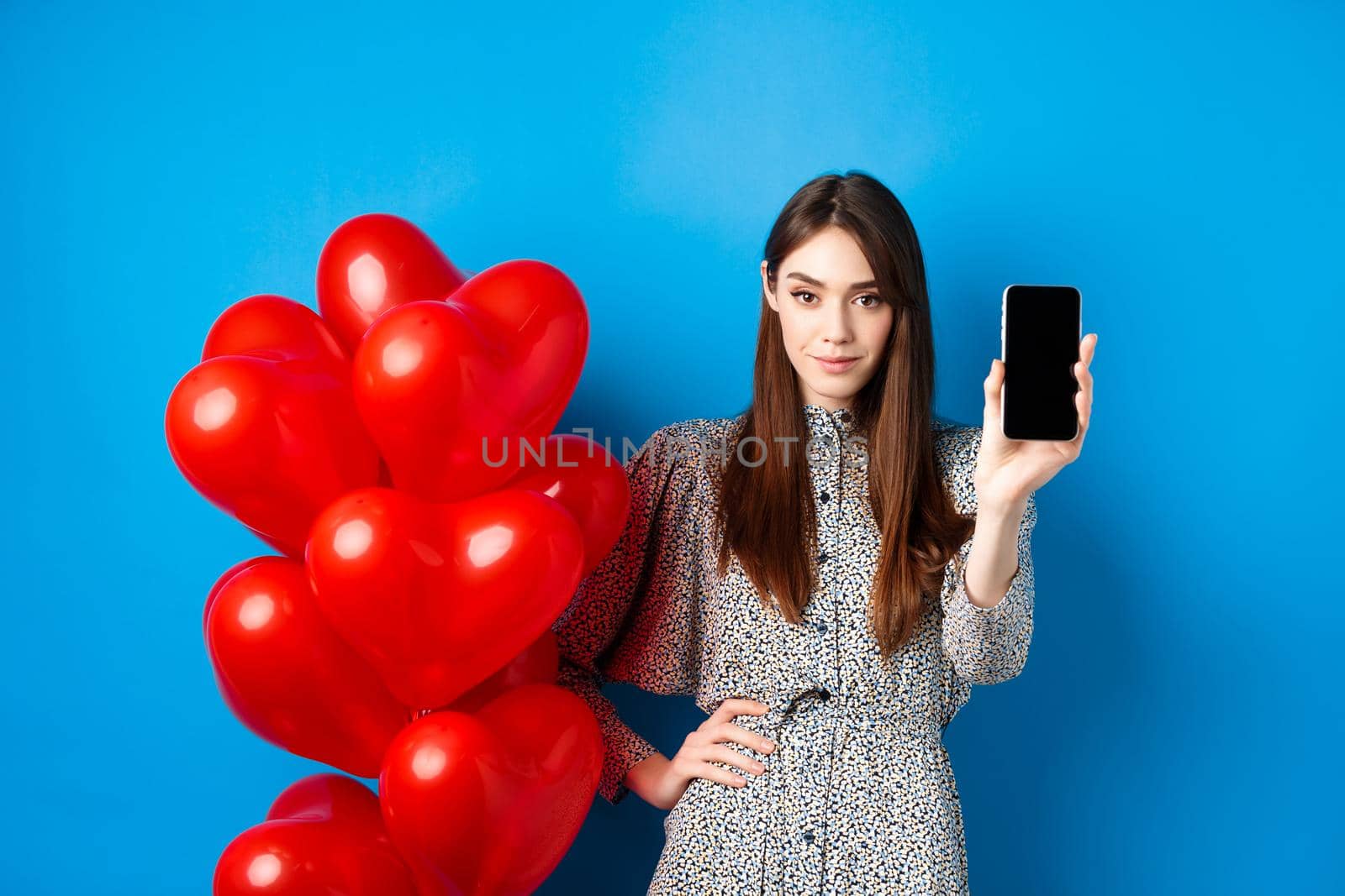 Valentines day. Beautiful young woman with makeup, wearing trendy dress, standing near red hearts balloons and showing empty mobile screen, blue background by Benzoix