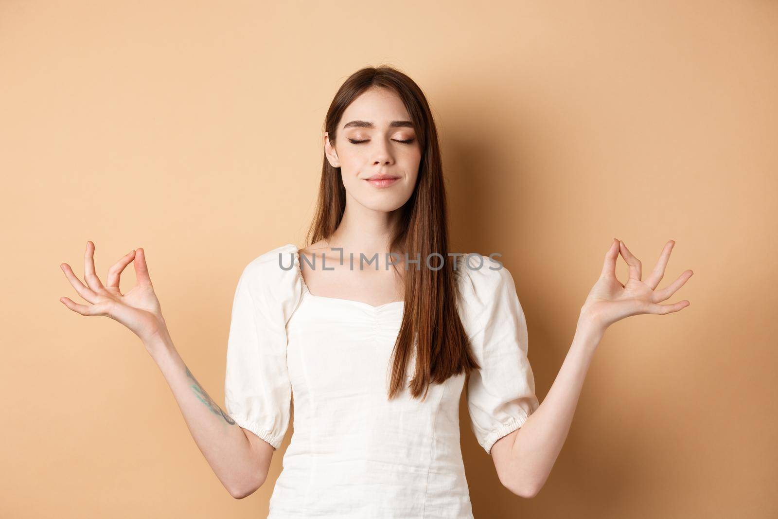 Relaxed young woman meditating, holding hands in yoga zen pose and close eyes, feel peace and smiling, standing on beige background.