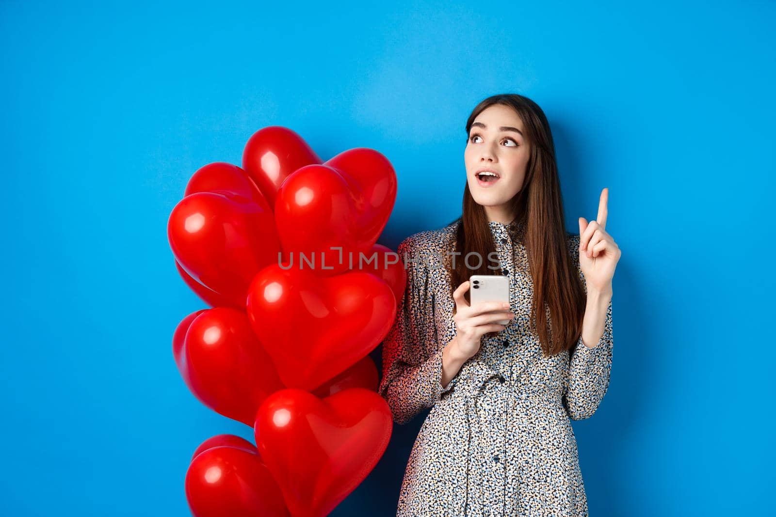 Valentines day. Image of romantic girl pitching an idea after using mobile phone, raising finger up and looking at empty space, standing near red heart balloons, blue background by Benzoix