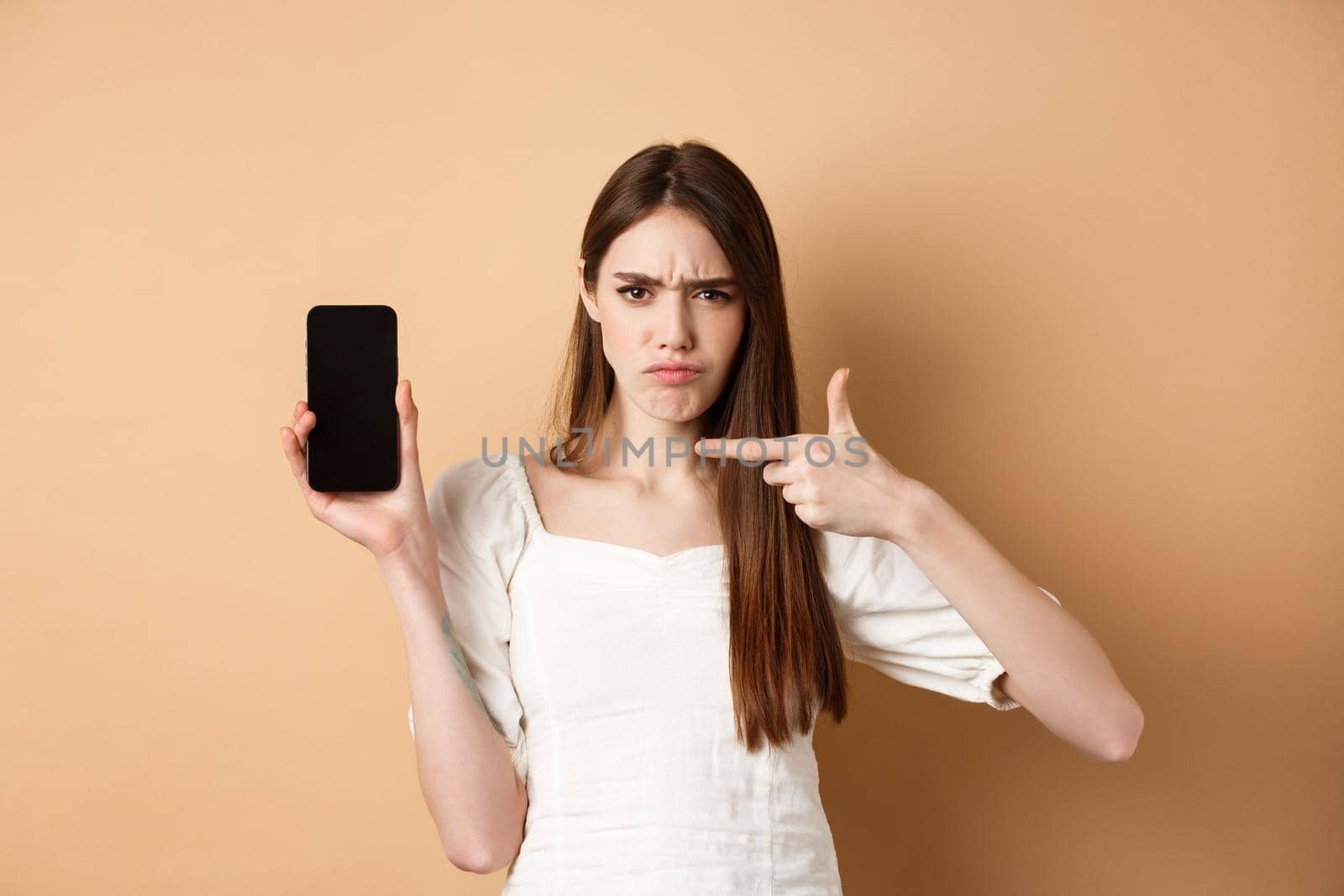 Disappointed frowning girl pointing at empty phone screen, complaining at online news, standing on beige background by Benzoix