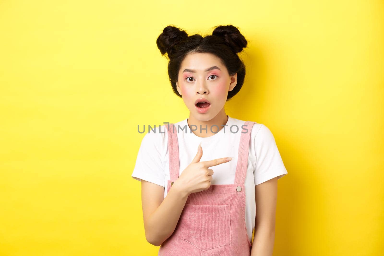 Shocked asian woman with glamour makeup, open mouth and pointing right at logo, showing advertisement, standing against yellow background.