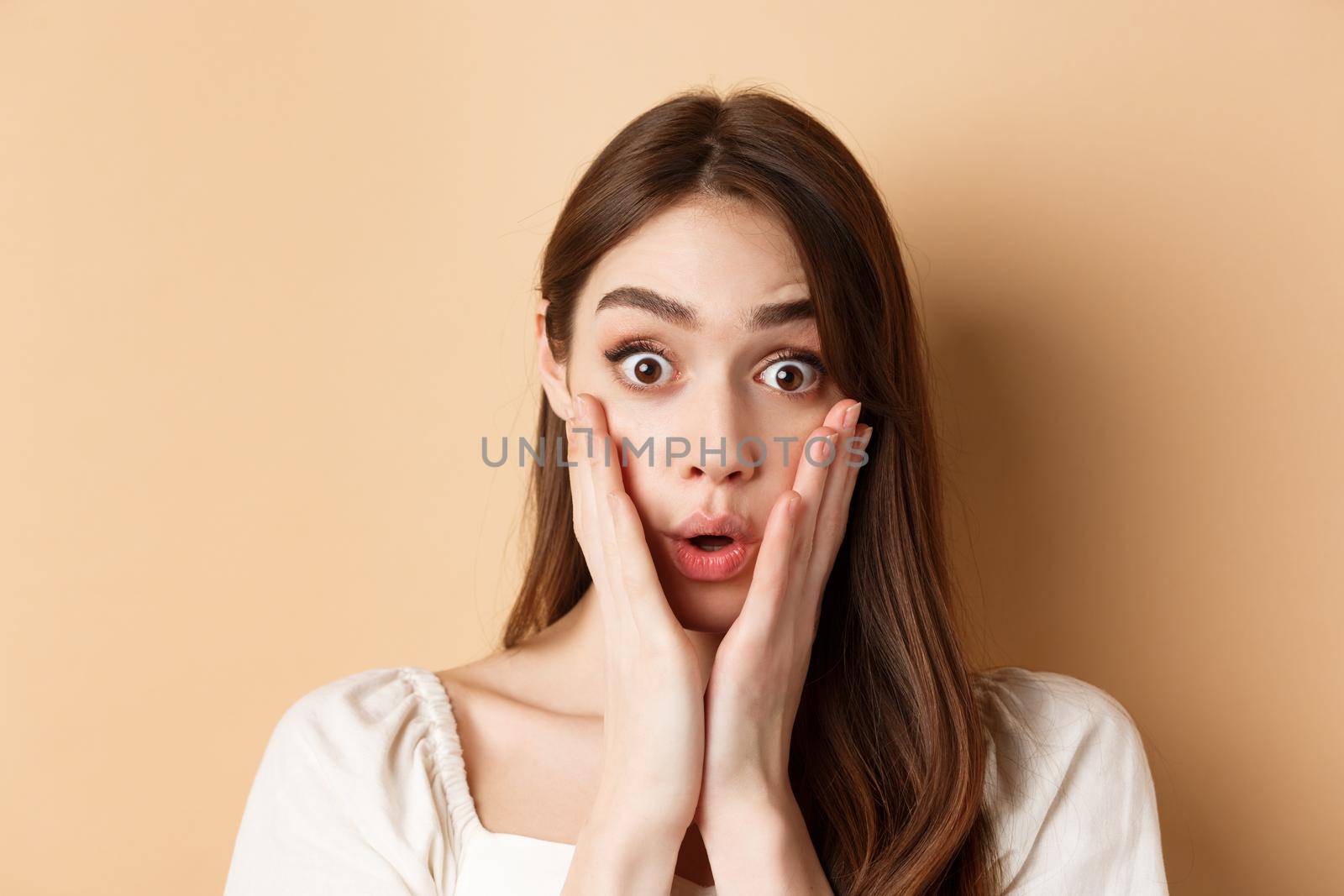 Face of surprised girl saying wow, touching cheeks and staring excited at camera, standing on beige background.