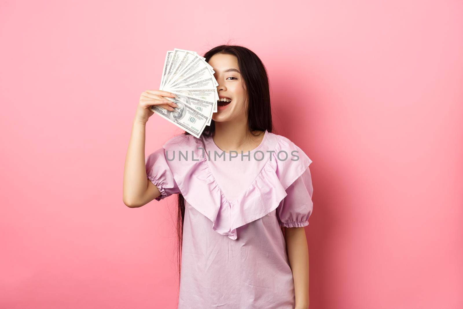 Happy rich asian woman showing money and smiling, shopping with cash, standing in dress against pink background.