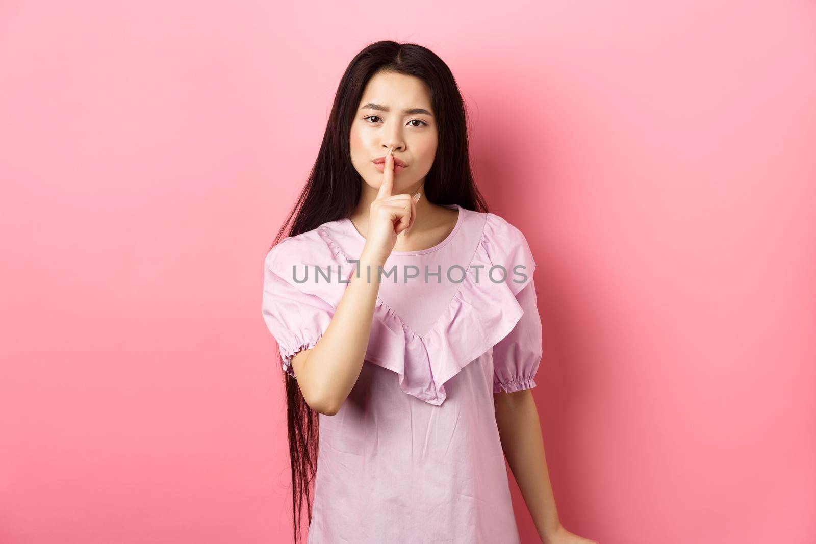 Cute asian girl tell to be quiet, scolding loud person, showing shush sign with finger pressed to lips, standing in dress on pink background.