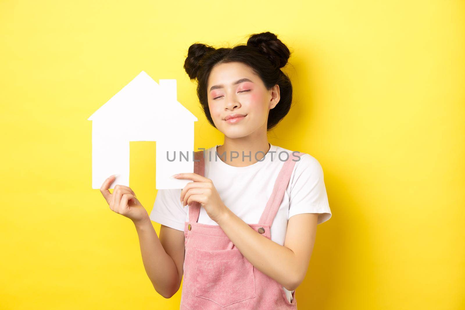 Real estate and family concept. Dreamy smiling asian woman with bright makeup, showing paper house cutout with closed eyes, daydreaming about buying property, yellow background by Benzoix