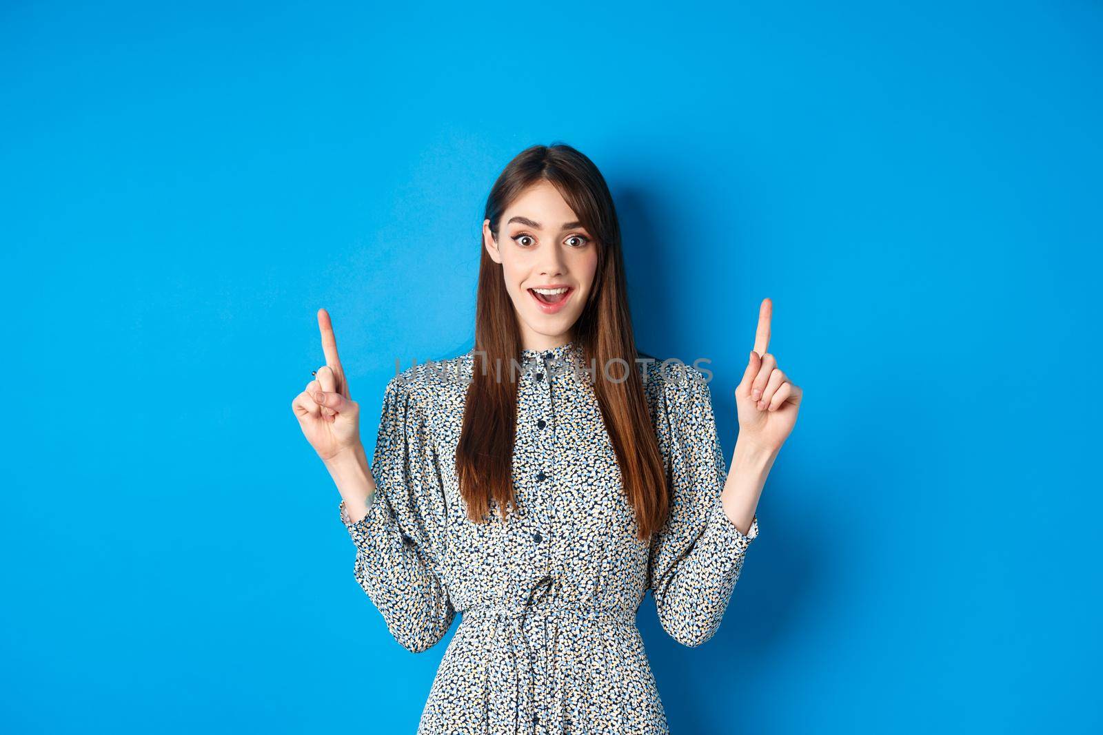 Excited and happy young woman in dress checking out special offer, pointing fingers up and smiling, standing against blue background by Benzoix