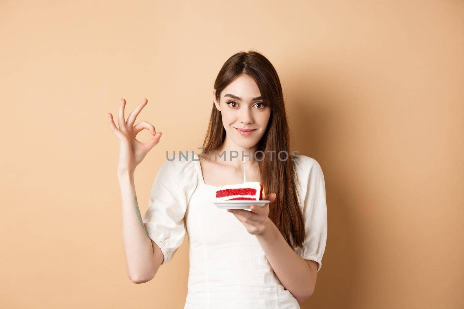 Pretty birthday girl holding cake and showing OK gesture, smiling pleased, enjoying bday celebration, beige background by Benzoix