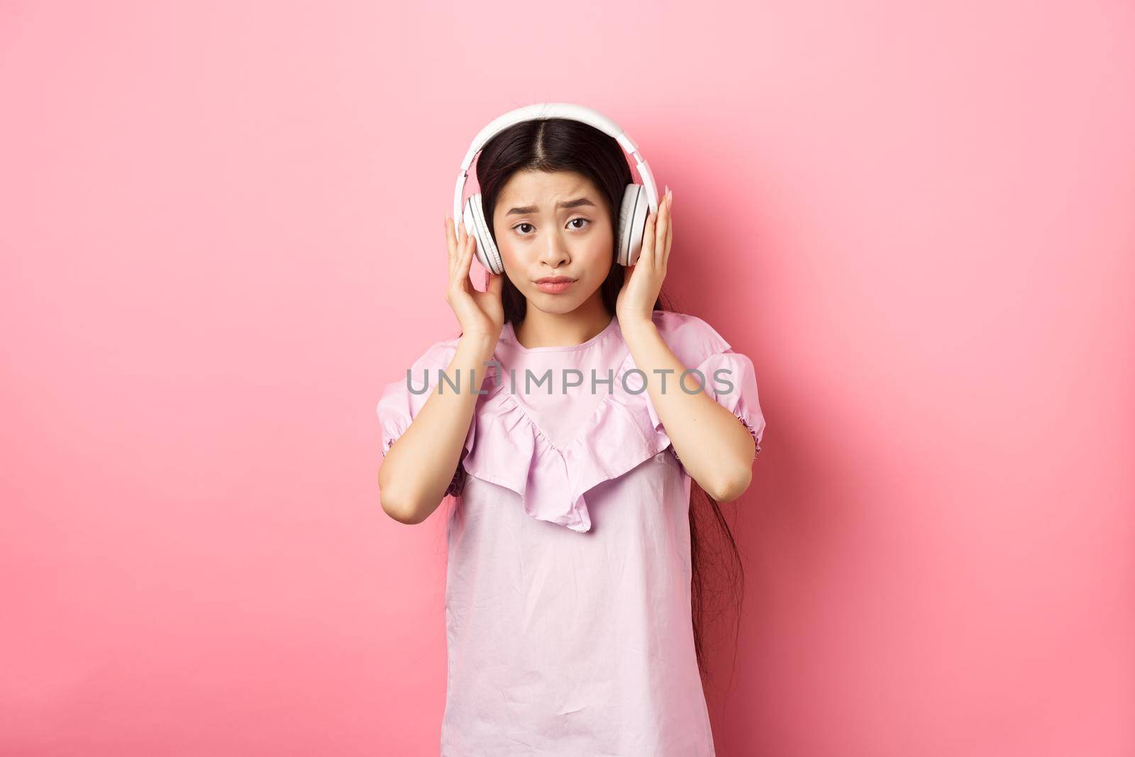 Skeptical girl unamused with song, listening music in headphones and frowning displeased, standing against pink background.