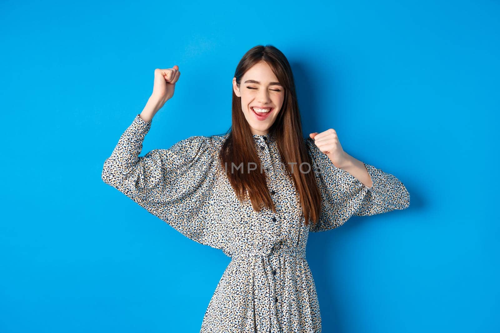 Excited smiling girl showing tongue and dancing with raised hands, celebrating victory or achievement, winning prize, standing happy on blue background.