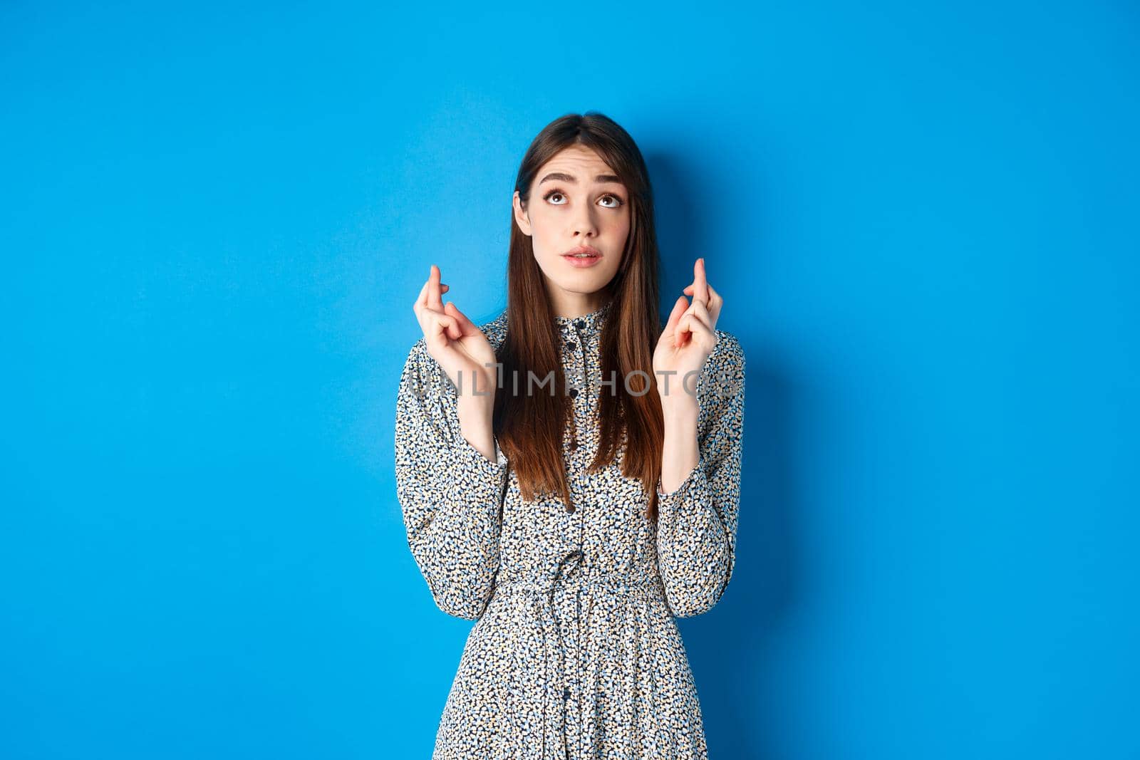 Hopeful cute young woman in dress with natural hair, looking up and cross fingers good luck, waiting for dream come true, praying or begging god, blue background.