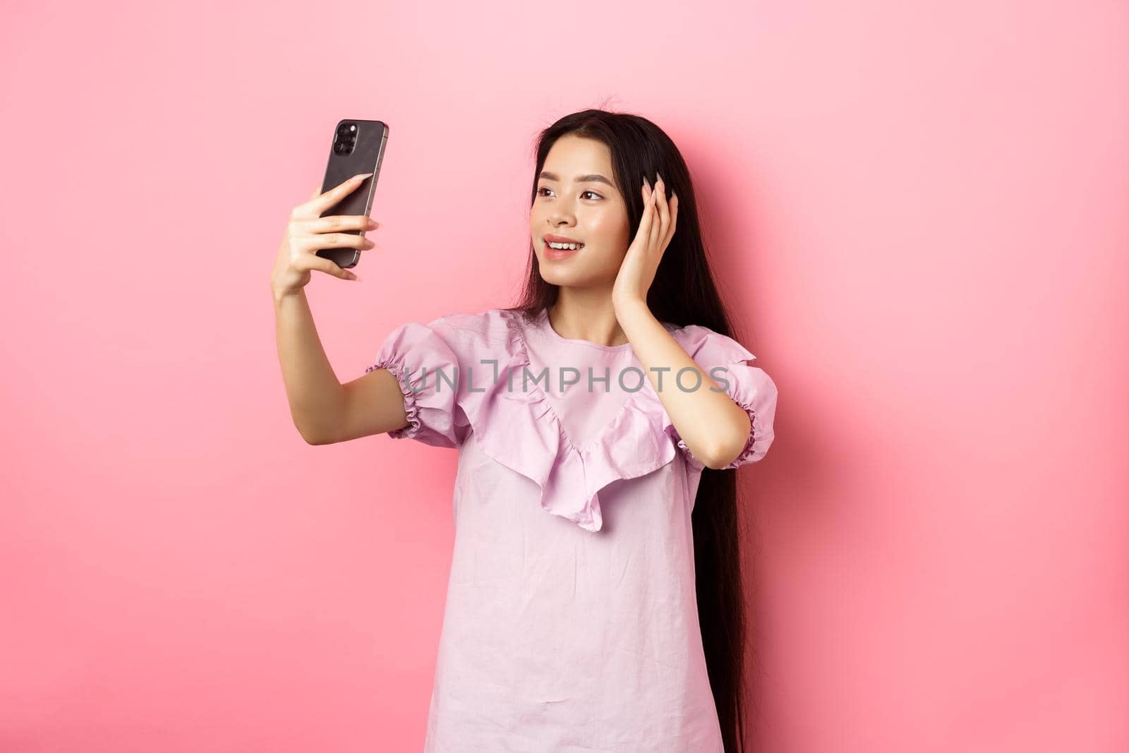 Stylish asian girl blogger taking selfie on mobile phone, posing for smartphone photo, standing in dress against pink background.