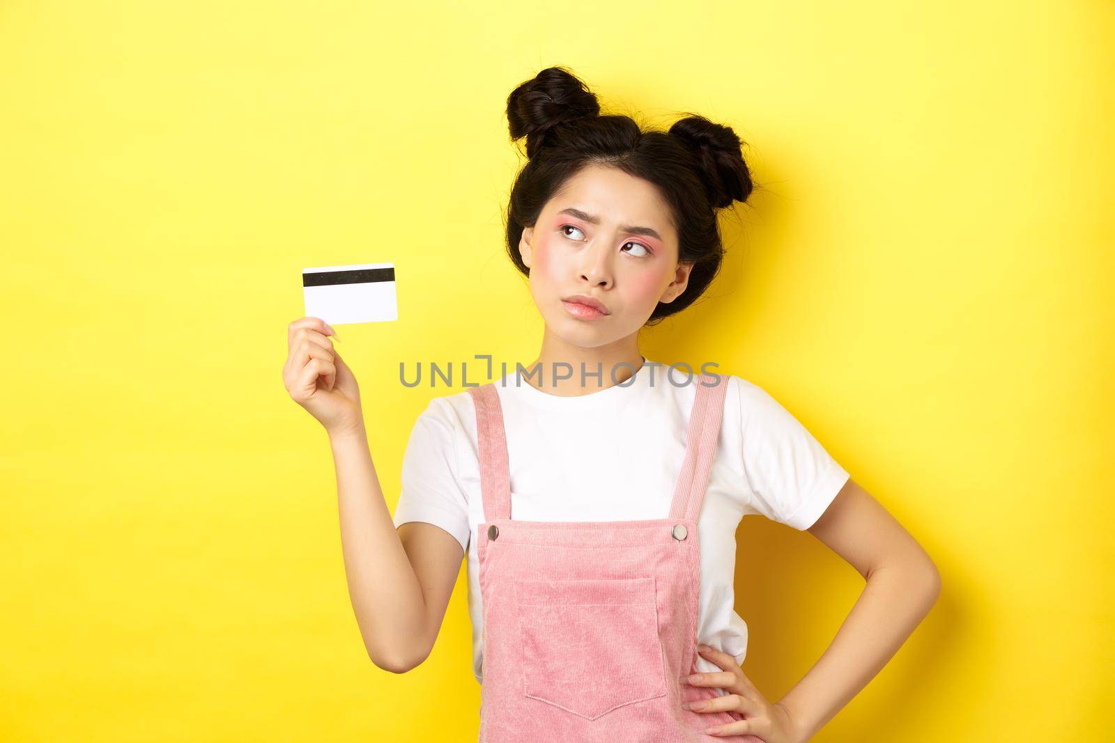 Shopping. Thoughtful asian woman with glamour makeup, holding plastic credit card and looking aside pensive, make decision, yellow background.
