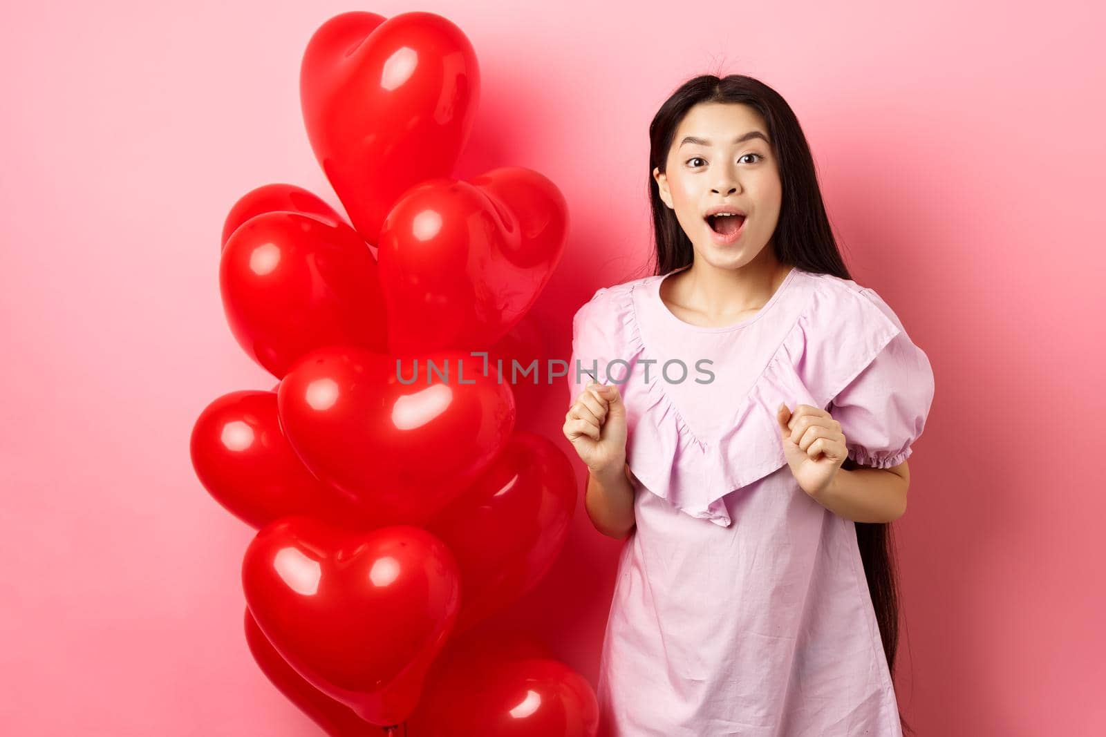 Excited and surprised teenage korean girl open mouth amazed, receive surprise gift on valentines day, looking wondered, standing near heart balloons, pink background.